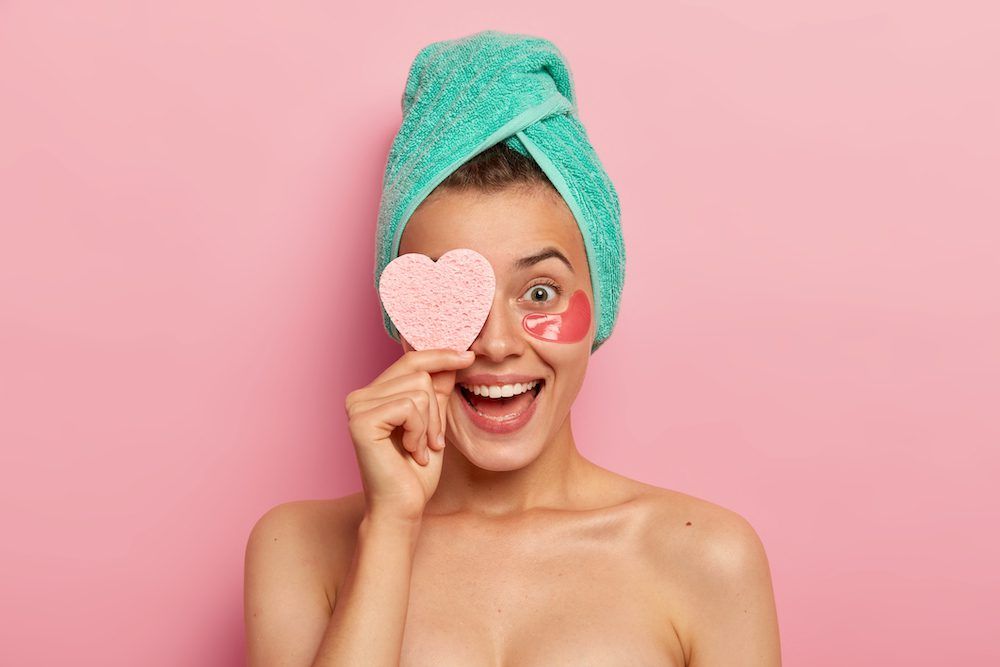 Portrait of overjoyed funny woman keeps cosmetic sponge on eye, laughs sincerely, wears undereye patches, removes wrinkles, cares about skin, has natural beauty, isolated over pink background